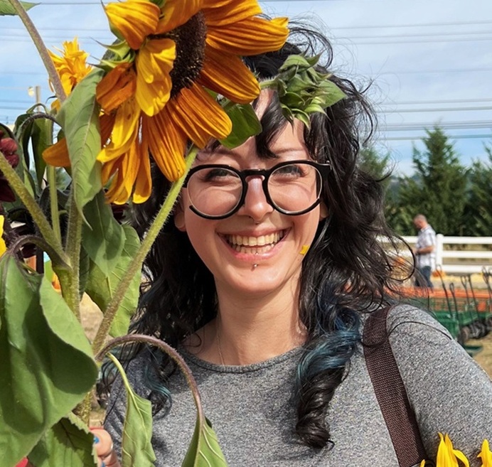 Sage smiling at the camera. She is wearing black-rimmed glasses and holding sunflowers.